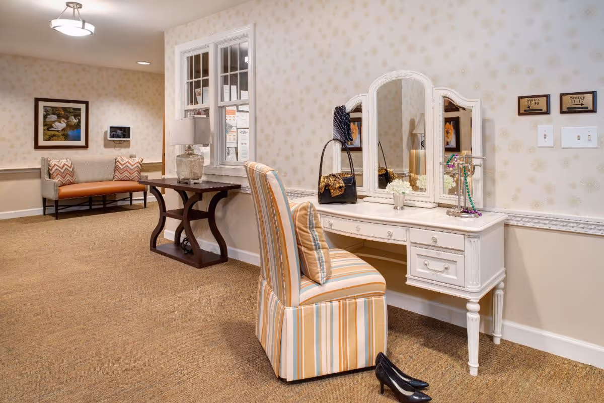 Bright assisted-living interior featuring a striped chair at a white vanity with mirror, a nearby side table and a seating bench along the wall.