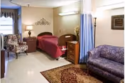 A furnished senior care bedroom with a single bed in a red bedspread, bedside tables and lamps, a floral armchair, a purple sofa, area rug, and a privacy curtain.