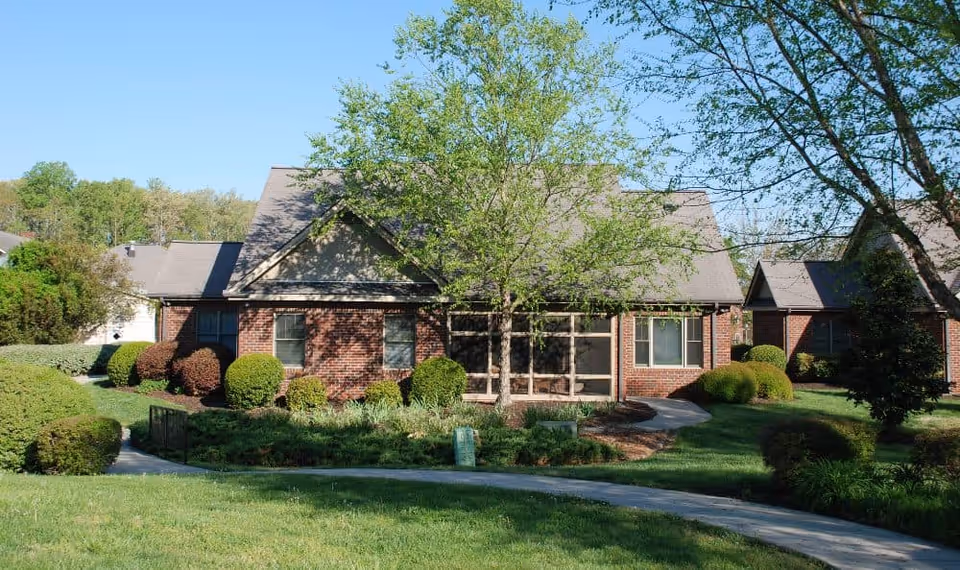 A single-story brick building with a gabled roof surrounded by well-maintained green lawns, bushes, and trees under a clear blue sky.