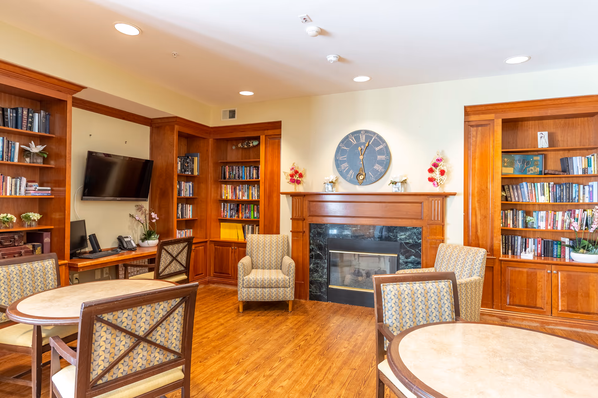 A cozy common area with wooden bookshelves filled with books, a fireplace with a large clock above it, and several upholstered chairs and round tables arranged for seating. A wall-mounted TV and a desk with a phone and computer are visible in the corner.