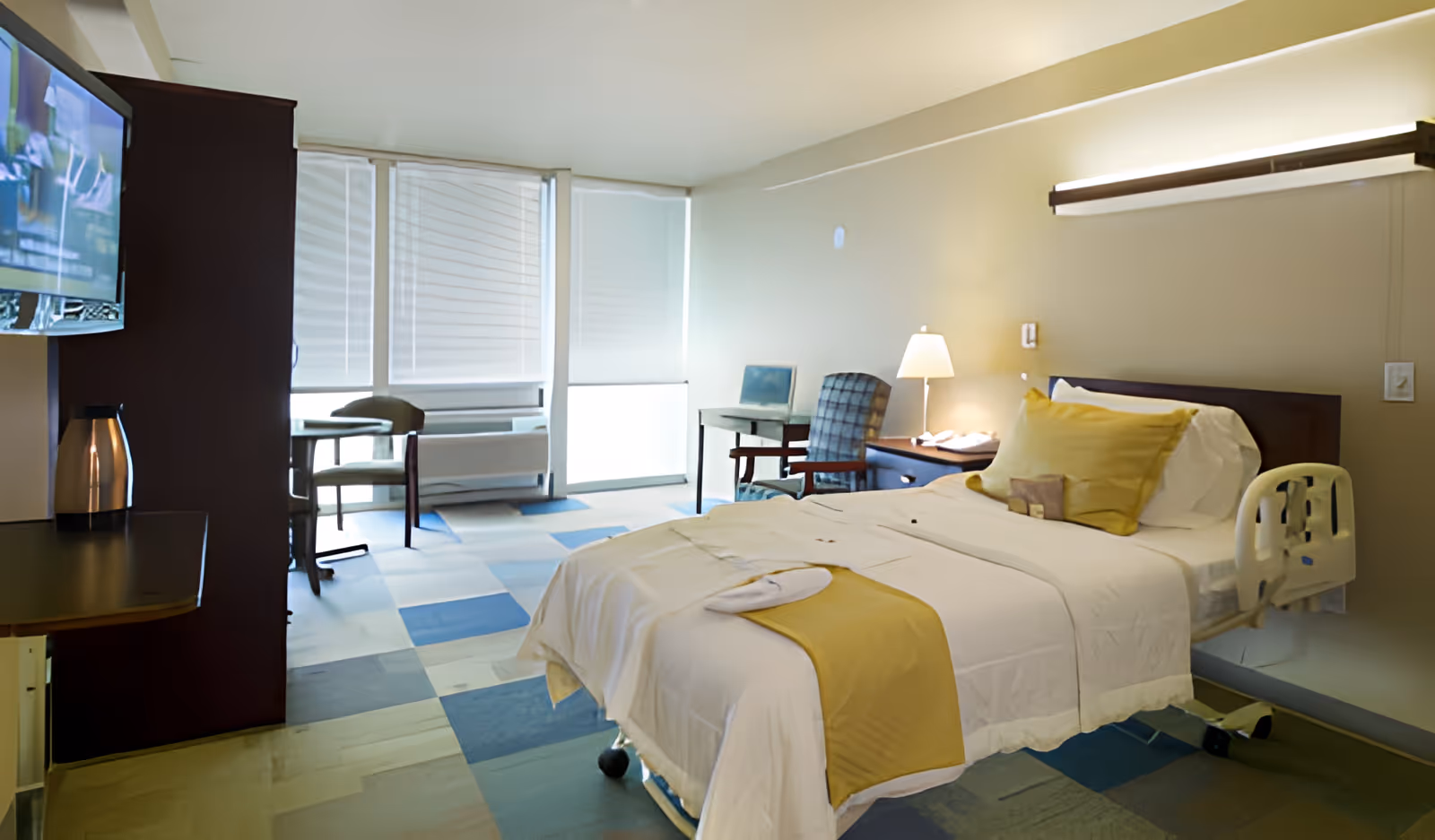 Well-lit patient bedroom with a single bed, bedside lamp and table, chairs, TV, and large windows.