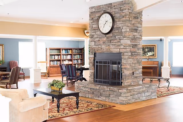 A spacious living area with a large stone fireplace in the center featuring a clock above it. The room has wooden floors, a beige armchair, a dark wooden coffee table with a plant, a patterned rug, bookshelves filled with books, and several chairs and tables arranged around the room. The walls are painted in light colors with large windows allowing natural light to fill the space.