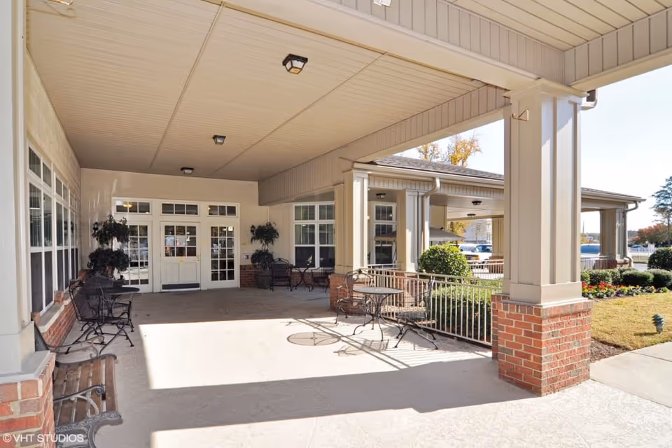 Covered outdoor patio area at Chesapeake Place Senior Living with metal tables and chairs, potted plants, and a bench. The area is adjacent to the building entrance with large windows and white double doors. The patio overlooks a landscaped garden with bushes and flowers.
