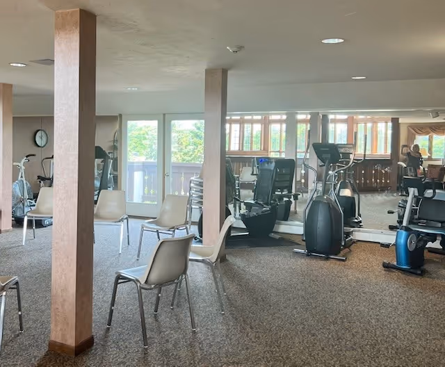 Interior view of a fitness room with exercise equipment including stationary bikes and elliptical machines. Several beige chairs are arranged in the room, and large windows allow natural light to enter. The room has carpeted flooring and light-colored walls with support columns.