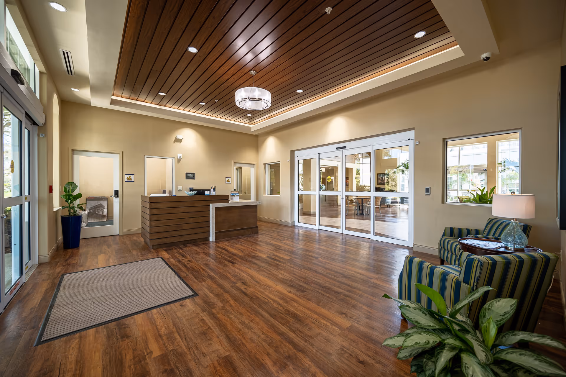 A spacious and well-lit reception area with wooden flooring and a wooden slatted ceiling. The reception desk is made of wood with a light-colored countertop. To the right, there are two striped armchairs with a small round table and a lamp between them. There are large glass sliding doors leading to another room with tables and chairs. Several plants are placed around the room, adding greenery to the space.