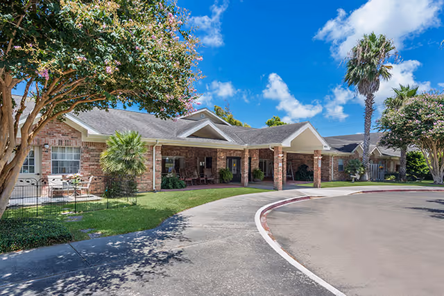 Exterior view of a single-story brick building with a covered entrance and a curved driveway. The building is surrounded by green grass, palm trees, and flowering trees under a bright blue sky with some clouds.