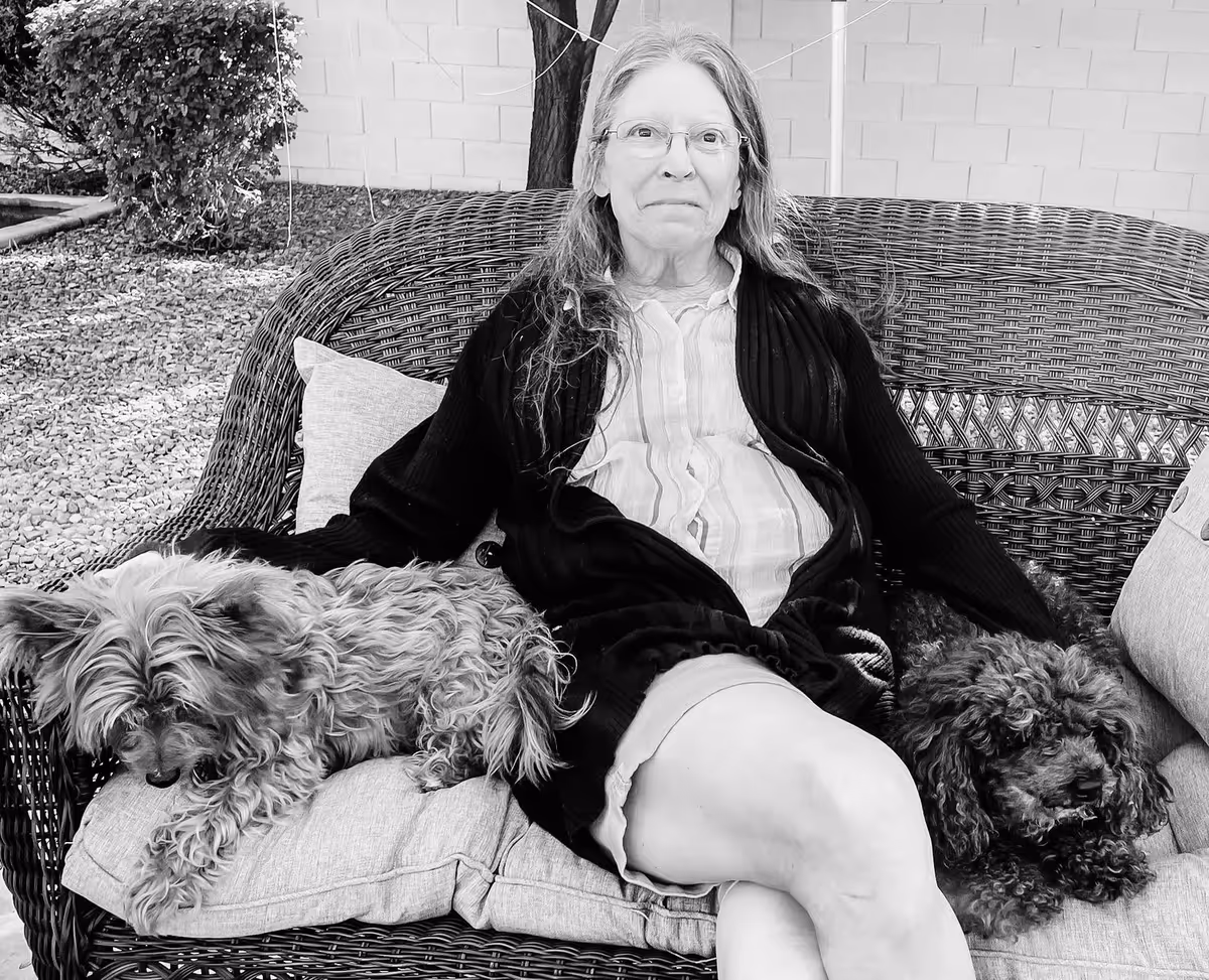 An elderly woman with glasses sitting on a wicker outdoor sofa with cushions, accompanied by two small dogs, one on each side. The setting appears to be an outdoor patio area with gravel ground and a tree in the background.