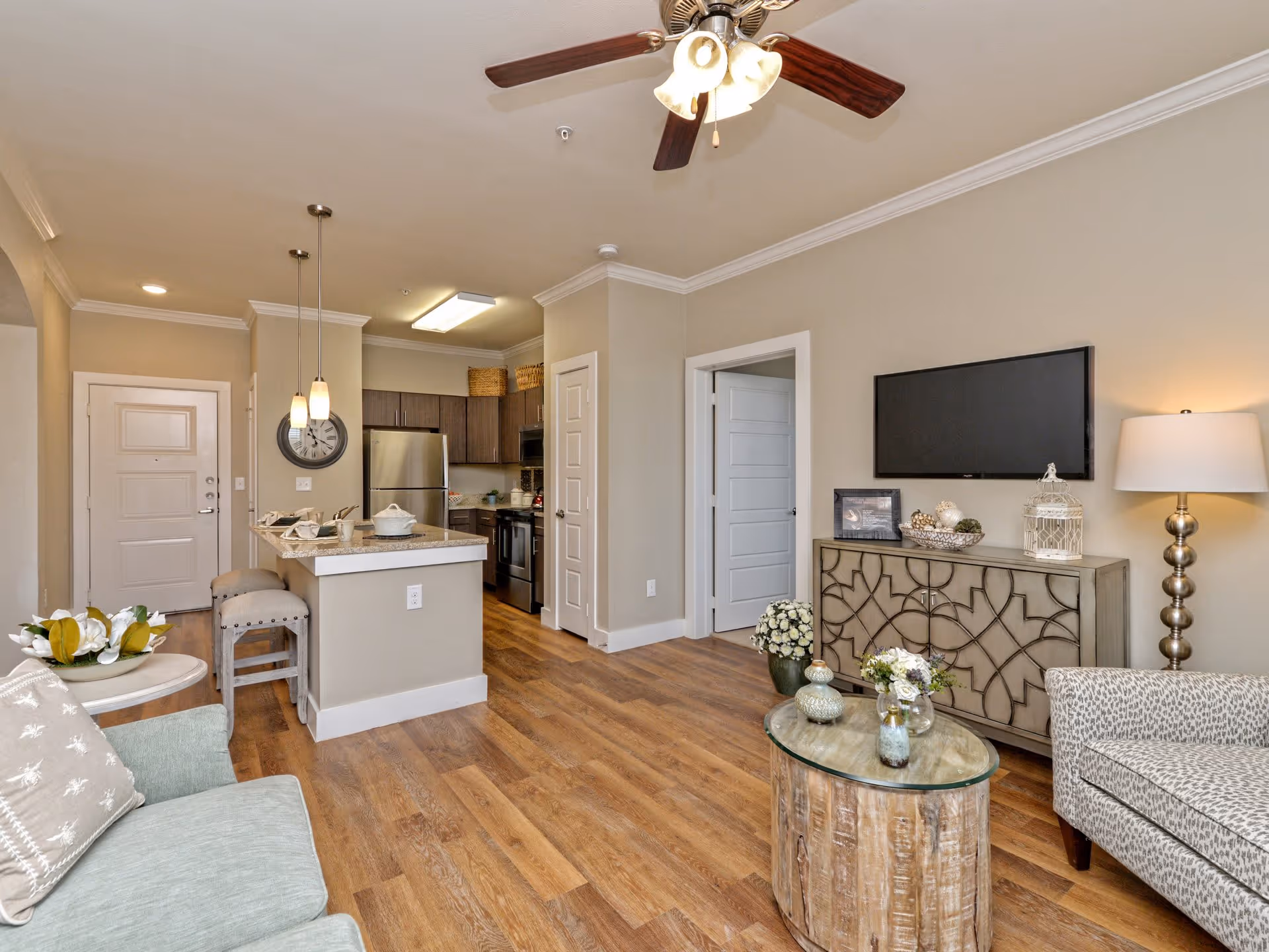 A modern senior living apartment interior featuring a living room and kitchen area. The living room has a light green sofa with a decorative pillow, a round glass-top wooden coffee table with small vases, a patterned armchair, a floor lamp, and a wall-mounted flat-screen TV above a decorative cabinet. The kitchen area includes a breakfast bar with two stools, pendant lights, stainless steel refrigerator, stove, and wooden cabinets. The flooring is wood, and the walls are painted beige with white trim. A ceiling fan with lights is visible above.