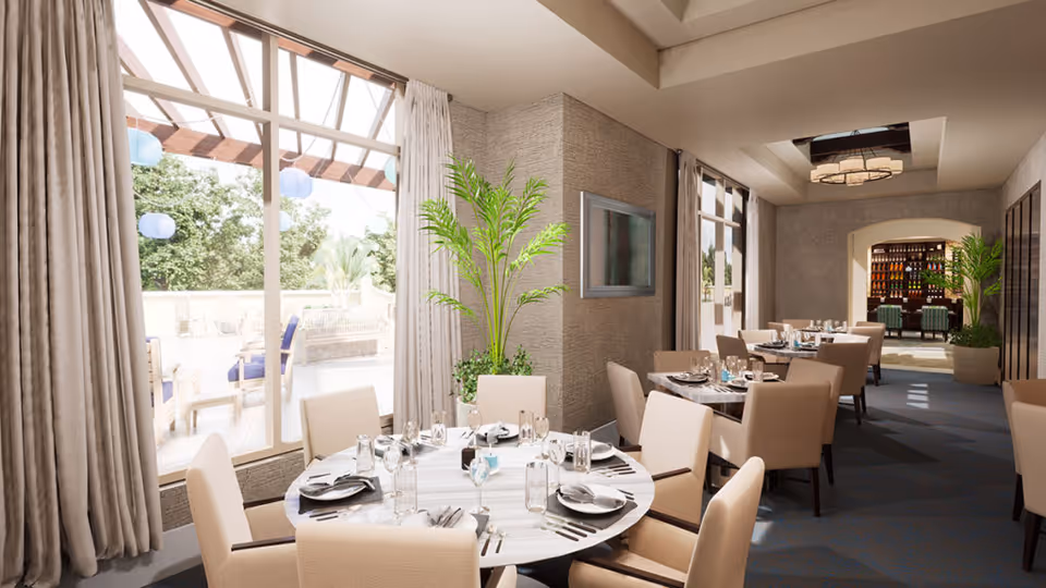A bright dining room with several tables set for a meal, featuring beige upholstered chairs and white marble tabletops. Large windows with beige curtains allow natural light to fill the room, and there are green plants placed around for decoration. In the background, there is a bar area with bottles displayed on shelves and additional seating.