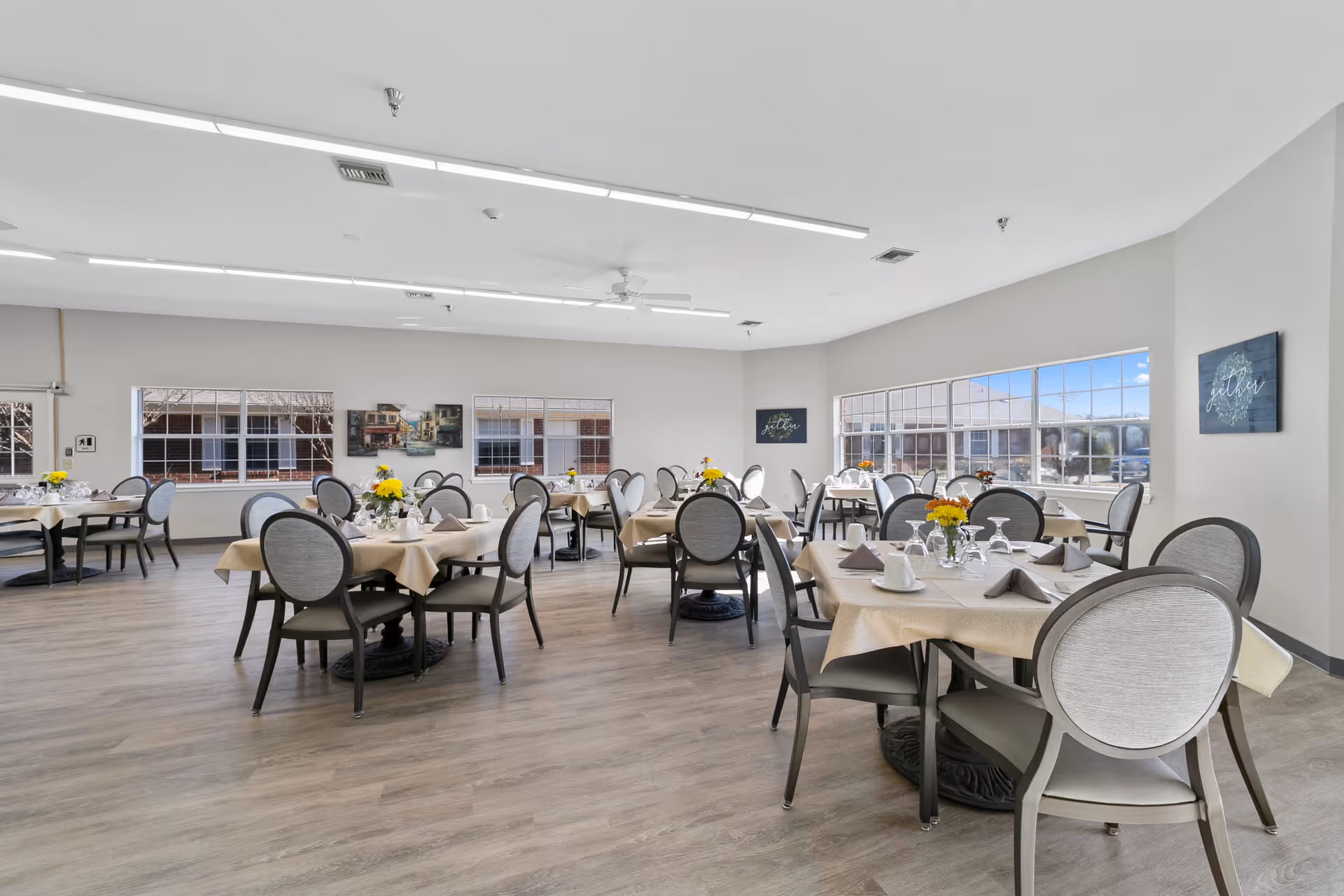 A bright dining room with multiple round tables covered with beige tablecloths, each set with plates, napkins, glasses, and small flower arrangements. The room has large windows letting in natural light, light-colored walls, and wood flooring.