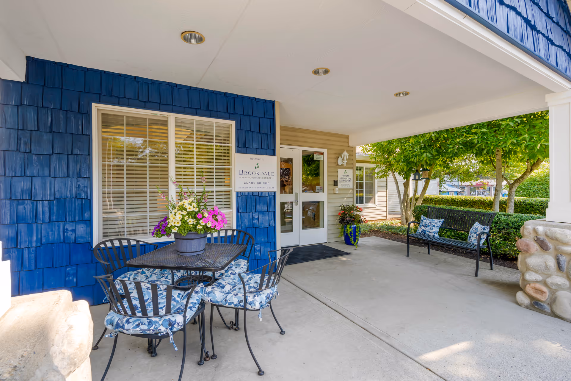 Covered outdoor seating area at the entrance of Brookdale Vancouver Stonebridge facility, featuring a black metal table with four chairs with blue floral cushions, a flower pot with colorful flowers on the table, a bench with two blue patterned pillows, and greenery in the background.