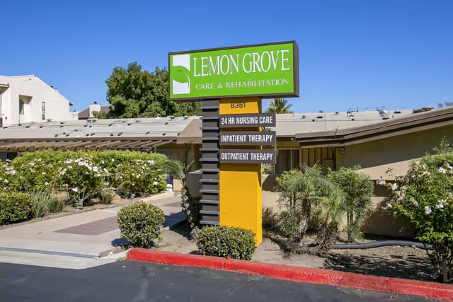 Exterior view of Lemon Grove Care & Rehabilitation Center with a large green and yellow sign displaying the facility name and services including 24 hr nursing care, inpatient therapy, and outpatient therapy. The building is surrounded by bushes and small palm trees under a clear blue sky.