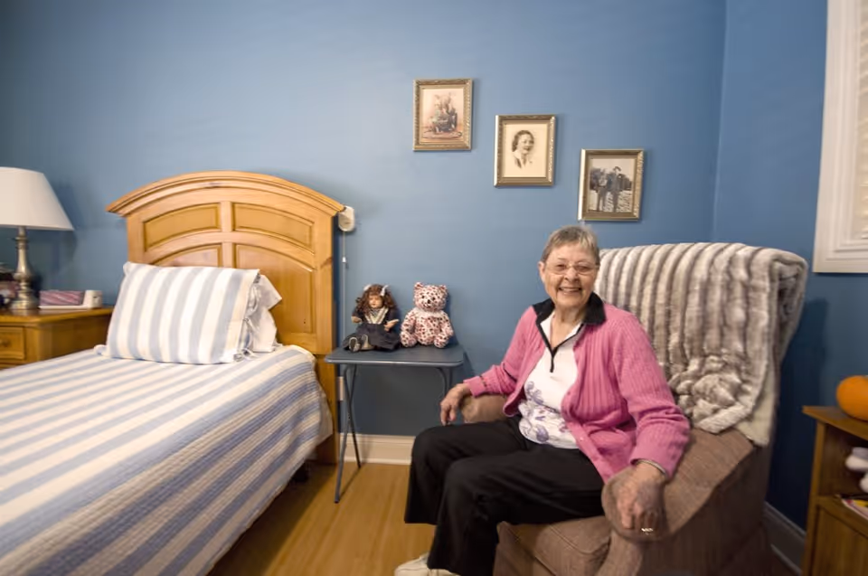 An elderly woman wearing a pink cardigan and glasses is sitting and smiling in a cozy room with blue walls. The room features a wooden bed with striped bedding, a nightstand with a lamp, a small table with two dolls, and three framed pictures hanging on the wall.