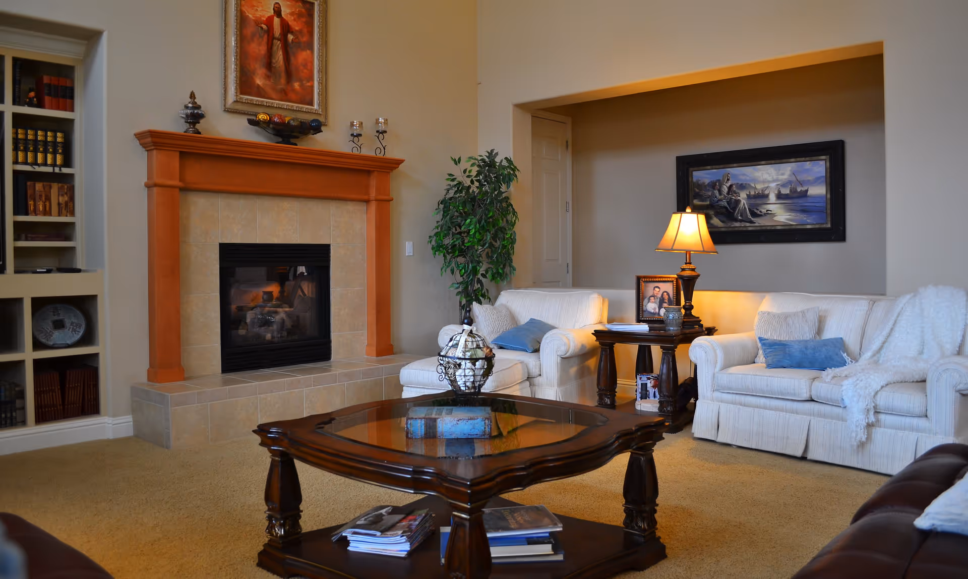 A cozy living room with a beige carpet, a fireplace with a wooden mantel, two white upholstered sofas with blue and beige pillows, a wooden coffee table with books and magazines, a side table with a lamp and framed photos, a green potted plant, and framed artwork on the walls.