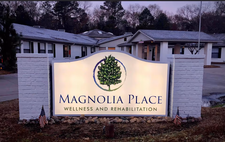 Illuminated outdoor sign for Magnolia Place Wellness and Rehabilitation in front of a single-story building with white brick pillars and American flags on either side of the sign during dusk.