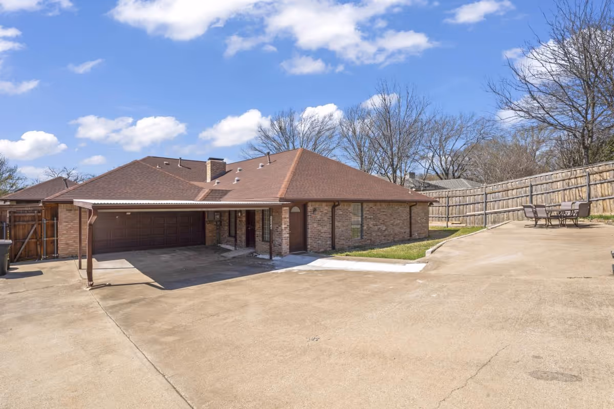 Exterior view of a single-story brick building with a brown roof under a blue sky with scattered clouds. The building has a covered carport and a driveway leading up to it. There is a wooden fence along the right side with an outdoor seating area consisting of a table and chairs.