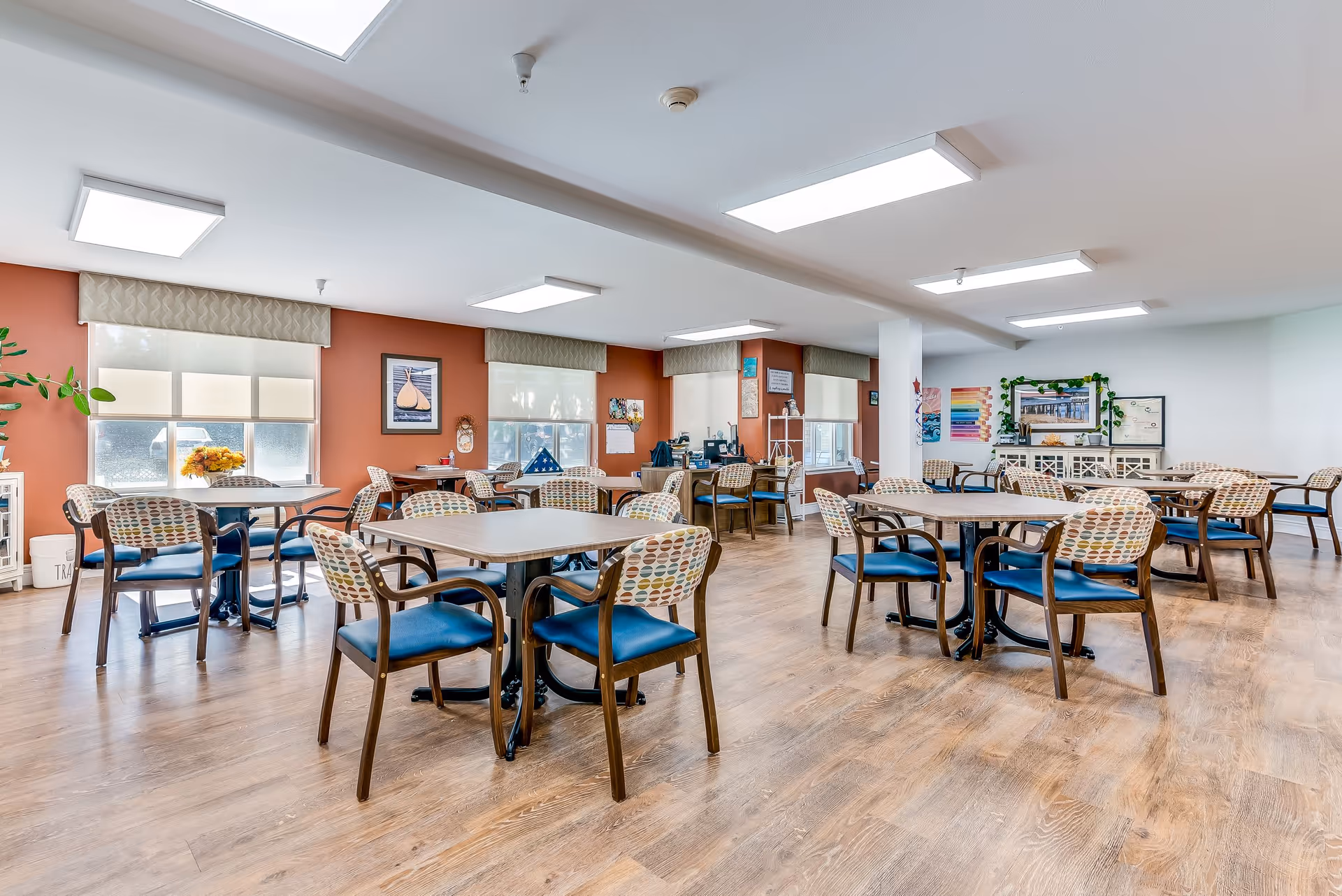 A bright and spacious dining room in a senior living facility with multiple square tables and chairs arranged neatly. The chairs have wooden frames with blue cushions and patterned backs. The room has large windows with beige valances, light wood flooring, and walls painted in a combination of white and warm orange. Various framed artworks and decorations adorn the walls, and there is a white sideboard with plants and certificates on the far wall.