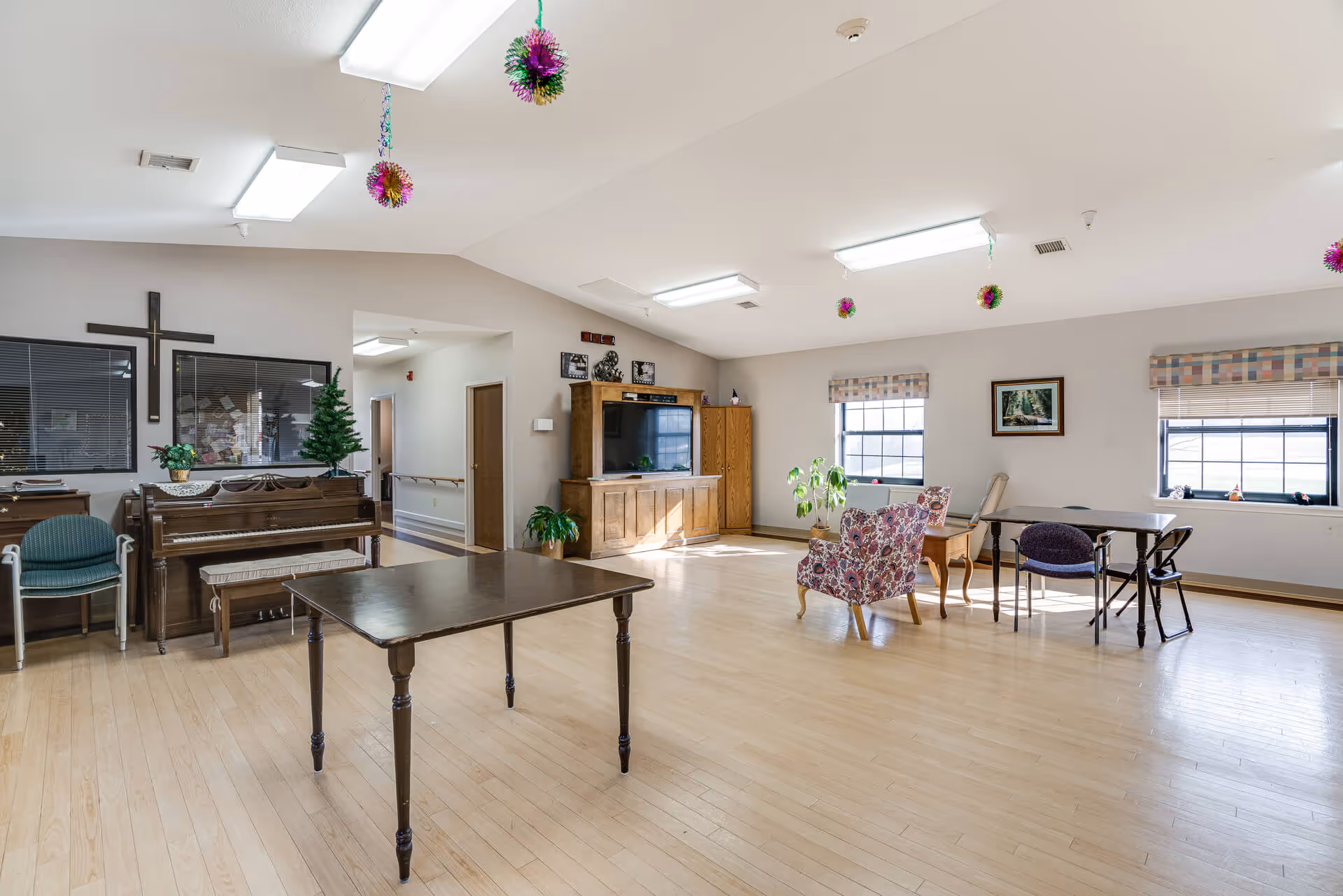 A spacious senior living common area with wooden floors, a piano against the wall with a bench, a large wooden table in the center, and several chairs including a floral armchair and a purple chair. There is a wooden entertainment center with a TV, two windows with patterned valances, a framed picture on the wall, and some hanging decorative ornaments from the ceiling. A cross is mounted on the wall above the piano.