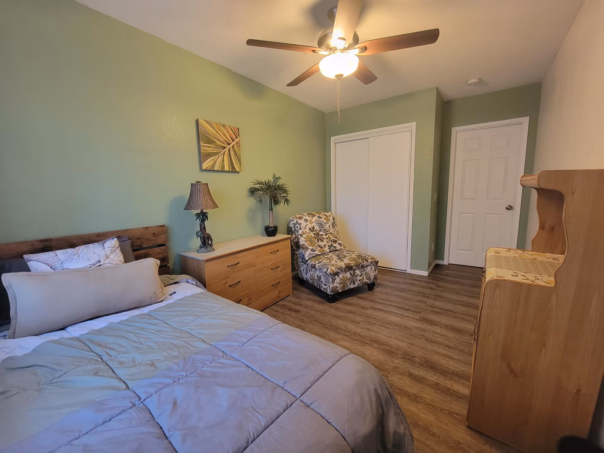 A tidy bedroom with a bed, wooden dresser, floral armchair, ceiling fan, and a green accent wall.