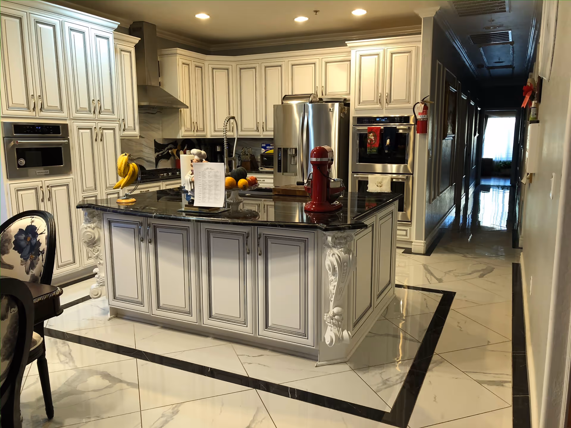 A spacious kitchen with white cabinetry and black granite countertops. The kitchen island features decorative molding and holds a red stand mixer, a fruit bowl with bananas and oranges, and a paper towel holder. Stainless steel appliances including a refrigerator, double oven, and microwave are visible. The floor is tiled with white tiles bordered by black. A hallway extends from the kitchen area with a fire extinguisher mounted on the wall.