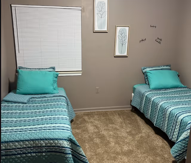 A small bedroom with two twin beds placed against opposite walls. Each bed has a turquoise and blue striped bedspread with matching pillows. There is a window with closed white blinds on the left wall and two framed botanical prints hanging on the center wall. Words like 'loving', 'patient', and 'gentle hands' are displayed on the right wall above one of the beds. The floor is carpeted in a light brown color.