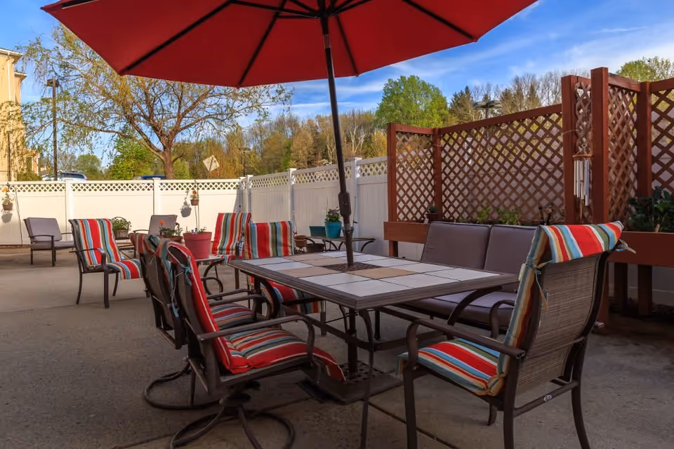 Outdoor patio area with a large table surrounded by chairs with colorful striped cushions. A red umbrella is open above the table. The patio is enclosed by a white fence and wooden lattice panels with some potted plants and greenery visible in the background under a blue sky.