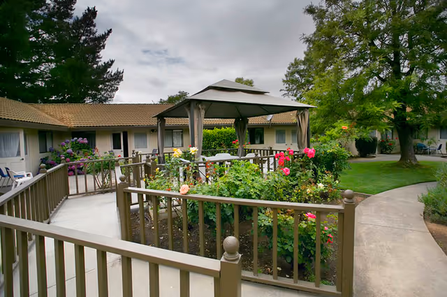 Outdoor garden area at Nazareth Rose Garden of Napa featuring a gazebo with curtains, surrounded by blooming flowers and greenery. The garden is enclosed by a brown wooden fence, with a paved walkway and single-story building with windows and doors in the background under a cloudy sky.