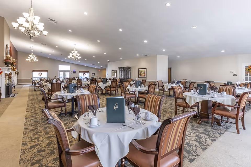 A spacious dining room with multiple round tables covered with white tablecloths, each set with glassware, napkins, and menus. The room features striped upholstered chairs, chandeliers hanging from the ceiling, and large windows letting in natural light. The decor is warm and inviting with framed artwork on the walls.