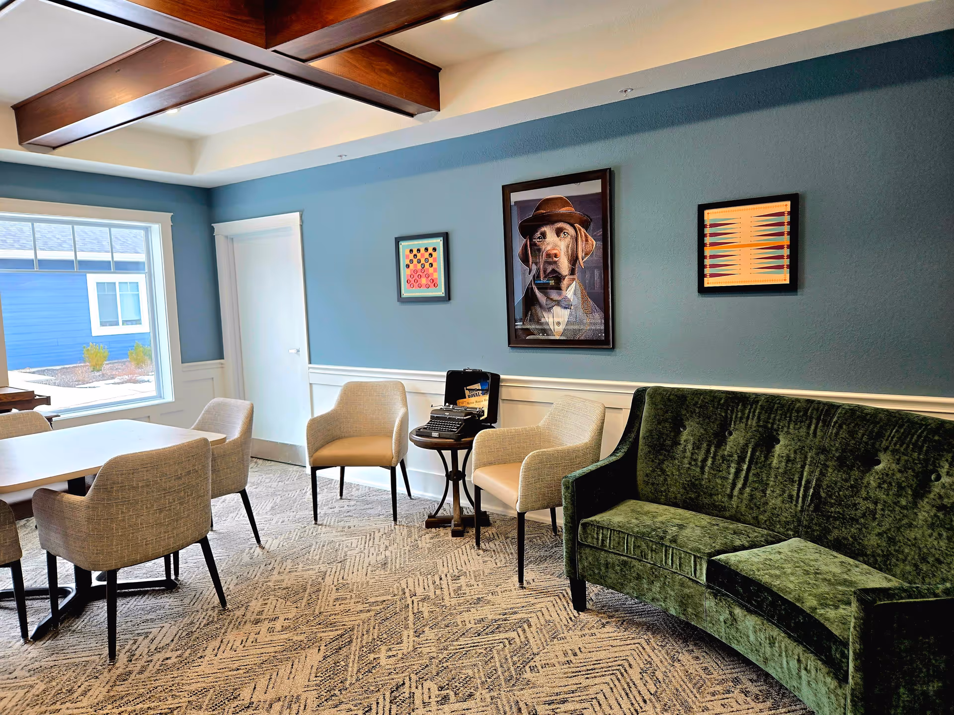 Bright senior living common room with a green velvet sofa, dining table and chairs, and framed artwork on teal walls.
