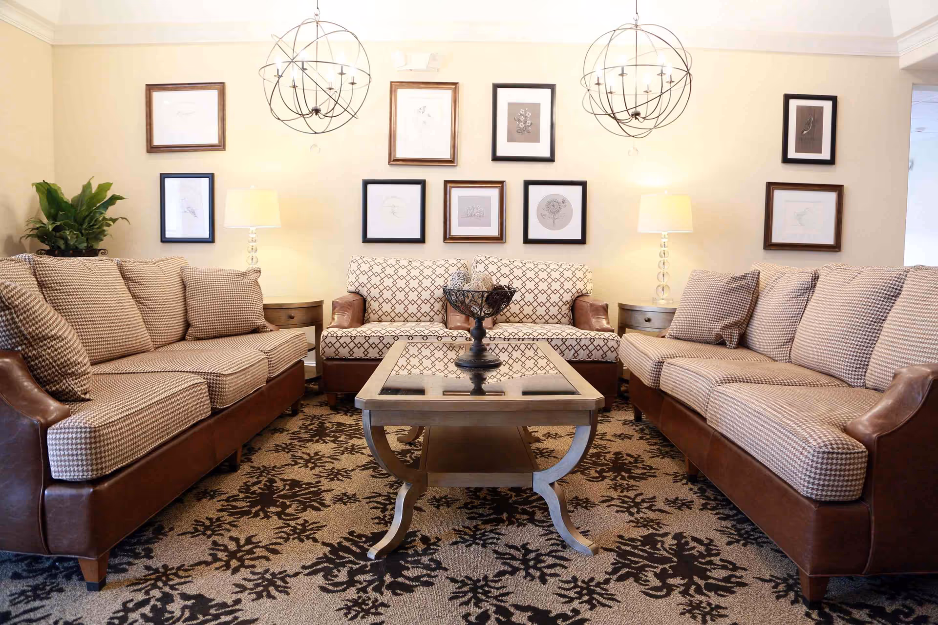 A cozy living room with two patterned sofas facing each other and a matching loveseat against the back wall. A glass-top coffee table with a decorative bowl is centered on a patterned carpet. The walls are adorned with framed artwork, and two modern spherical chandeliers hang from the ceiling. Two table lamps on side tables provide warm lighting, and a green plant is placed in the corner.