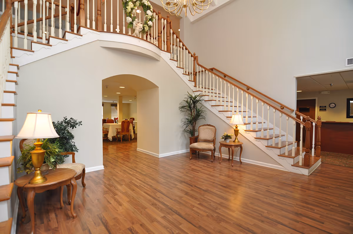 Interior view of a senior living facility lobby with a wooden staircase featuring white balusters and wooden handrails. The area has hardwood flooring, two small wooden tables with lamps and plants, and two upholstered chairs. In the background, a dining area with tables and chairs is visible through an arched doorway.