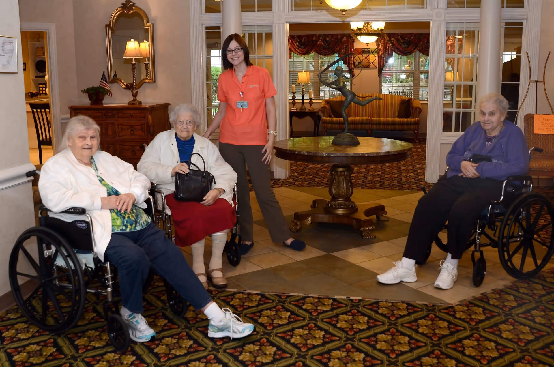 Three elderly women, two in wheelchairs and one standing, are gathered in a warmly decorated common area with patterned carpet and a round wooden table with a sculpture on it. The standing woman, wearing an orange shirt and glasses, smiles while posing with the seated women. The background shows a cozy sitting area with a striped couch, lamps, and large windows.