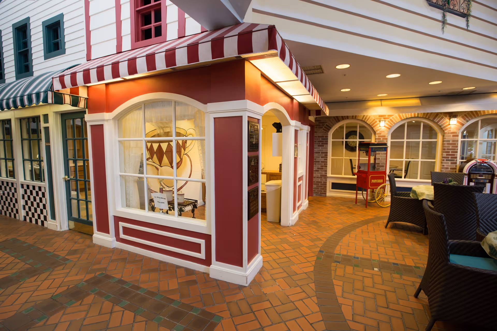 Indoor area designed to look like a small town street with storefront-style facades, including a red and white striped awning, a popcorn machine, wicker chairs, and tables with tablecloths on a brick floor.