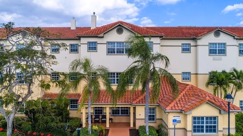 Exterior view of a multi-story senior living facility with beige walls and red tiled roof, surrounded by palm trees and landscaped greenery under a partly cloudy sky.
