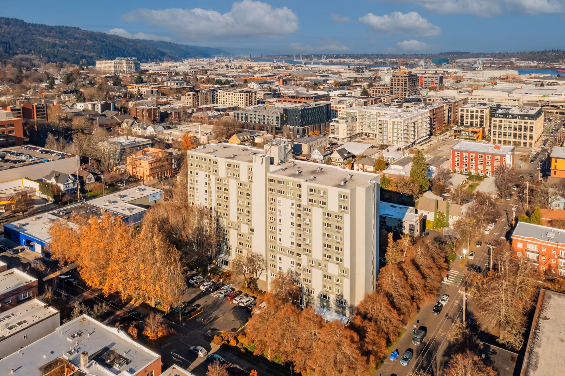 Aerial view of a cityscape featuring a tall, light-colored apartment building surrounded by trees with autumn foliage. The area includes various other buildings, streets with parked cars, and hills in the background under a partly cloudy sky.