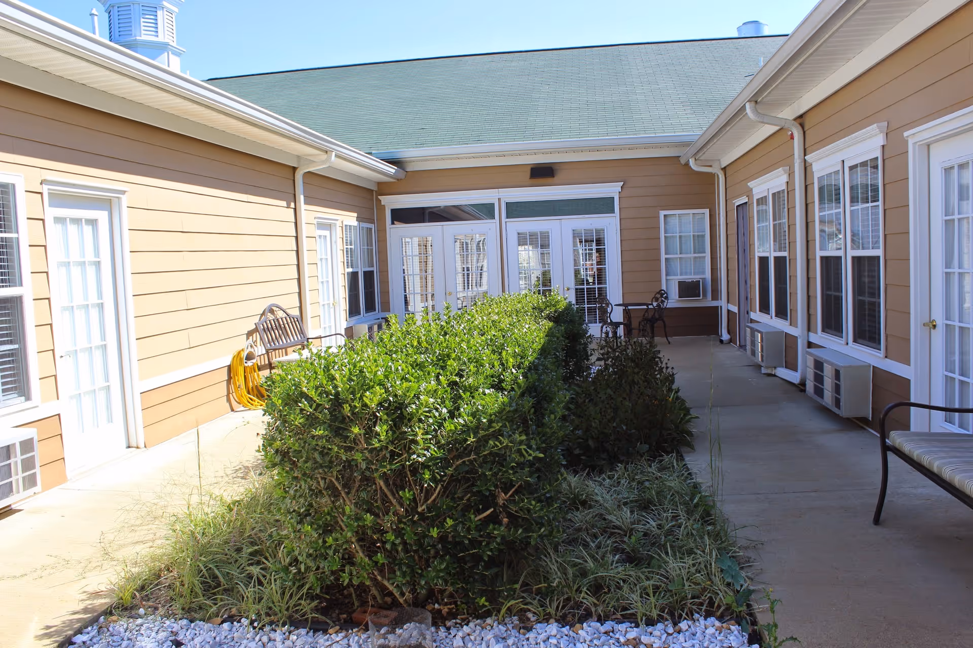 Outdoor courtyard area at SomerOak Senior Living with beige siding buildings surrounding a central garden with green bushes and plants. There are several windows and doors with white frames, a bench, and a small table with chairs on the concrete walkway.