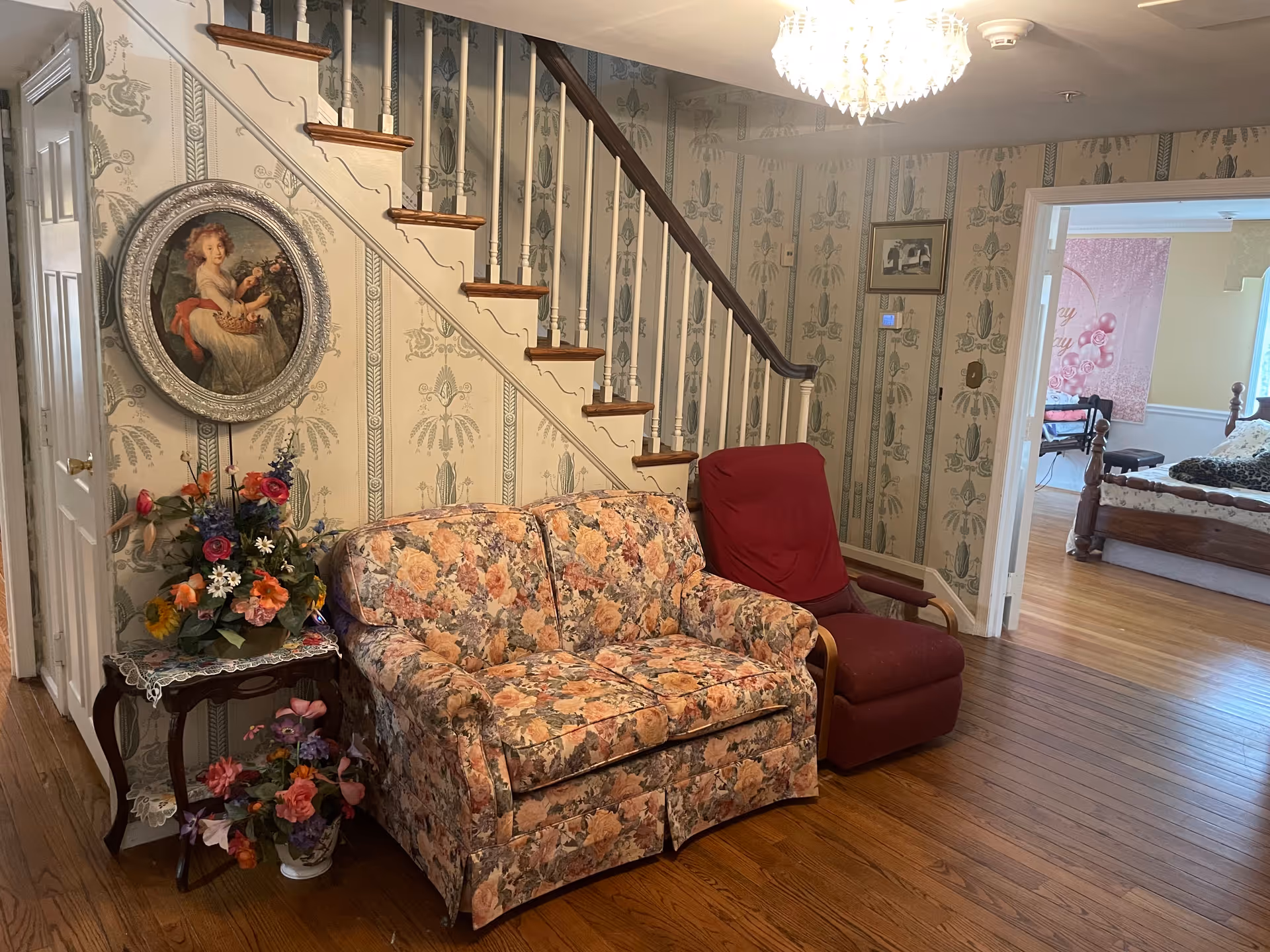 A cozy interior living area with floral wallpaper and wooden flooring. There is a floral-patterned loveseat and a red armchair placed near a staircase with white spindles and wooden handrail. A small wooden side table with a lace doily holds a colorful flower arrangement. A framed portrait of a young girl hangs on the wall above the loveseat. In the background, an open doorway reveals a bedroom with a wooden bed and a pink wall decoration.