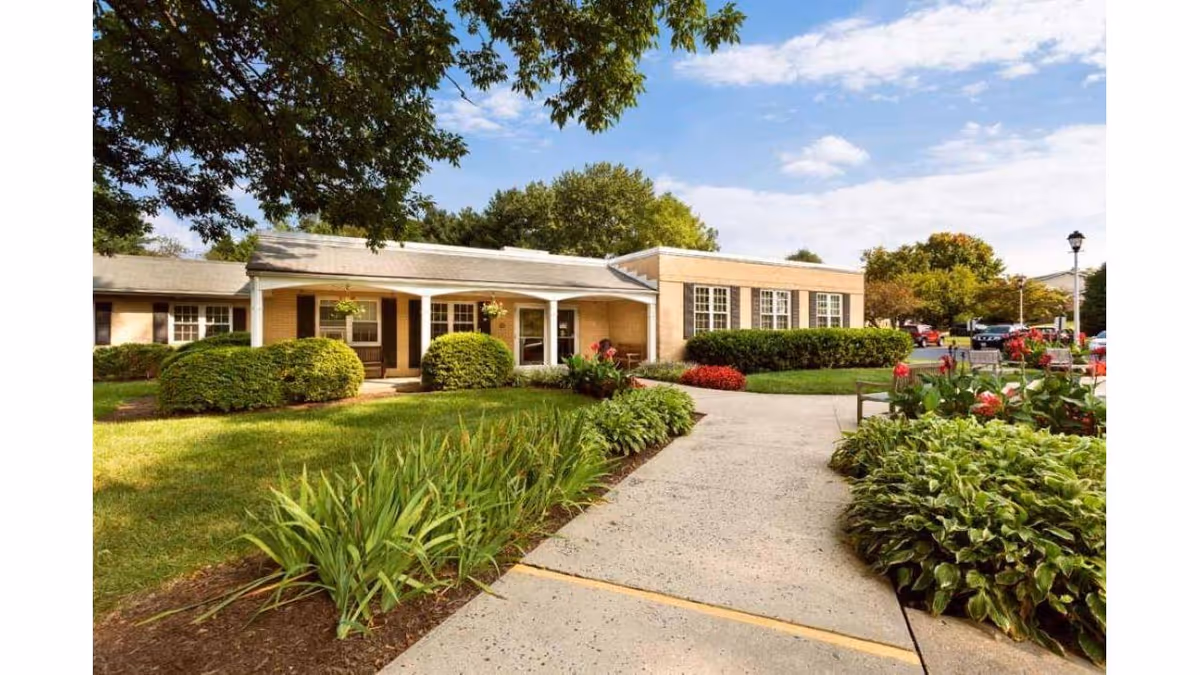 Exterior view of Sterling Care Bel Air facility showing a single-story building with beige walls and white trim, surrounded by well-maintained green bushes, plants, and trees under a partly cloudy sky. A concrete walkway leads to the entrance, and there are benches and a parking area visible in the background.