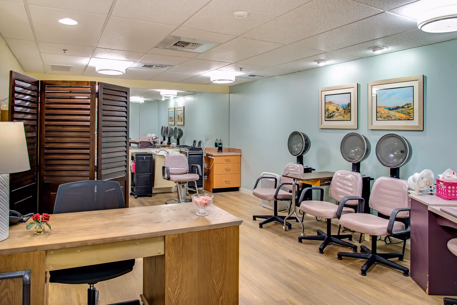 Interior view of a salon area in a senior living facility with several pink salon chairs and hair drying stations along the right wall. A wooden desk with a black office chair is in the foreground, and a large mirror covers the back wall reflecting the room. The walls are painted light blue and decorated with framed landscape paintings. The floor is wood, and the ceiling has recessed lighting.