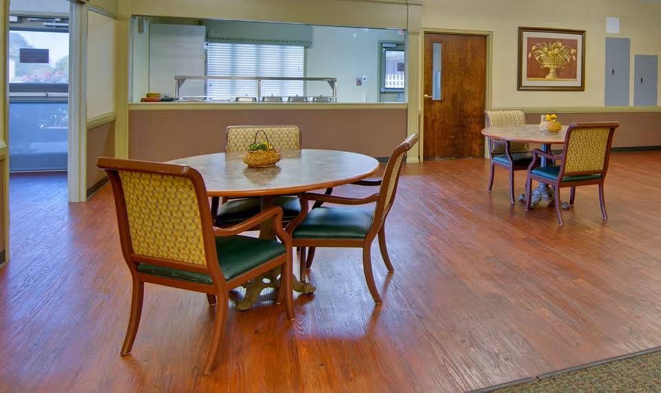 Dining area with round wooden tables and upholstered chairs on a wood floor in front of a serving window.