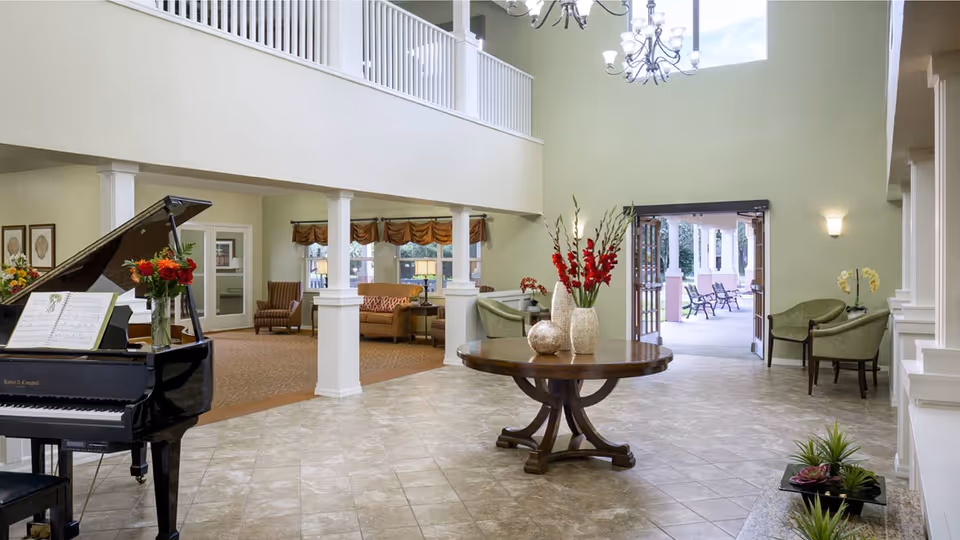 Spacious senior living facility common area with a grand piano on the left, a round wooden table with decorative vases and flowers in the center, and seating areas with chairs and sofas in the background. Large windows and an open doorway lead to an outdoor patio with benches. The room features high ceilings, chandeliers, and light green walls.