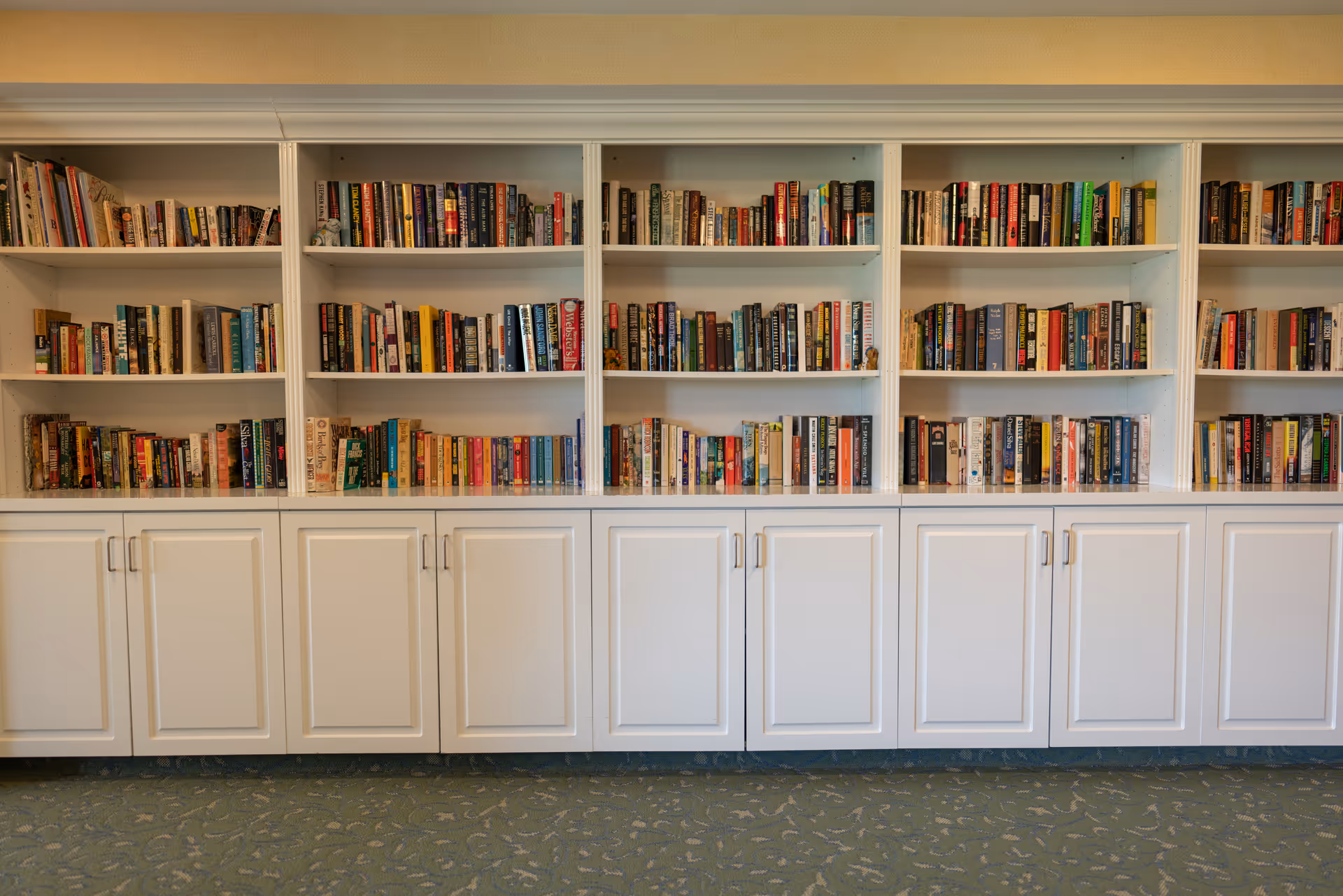 A large white bookshelf filled with numerous books arranged neatly on multiple shelves above white cabinets with closed doors, set against a wall with a patterned carpeted floor.