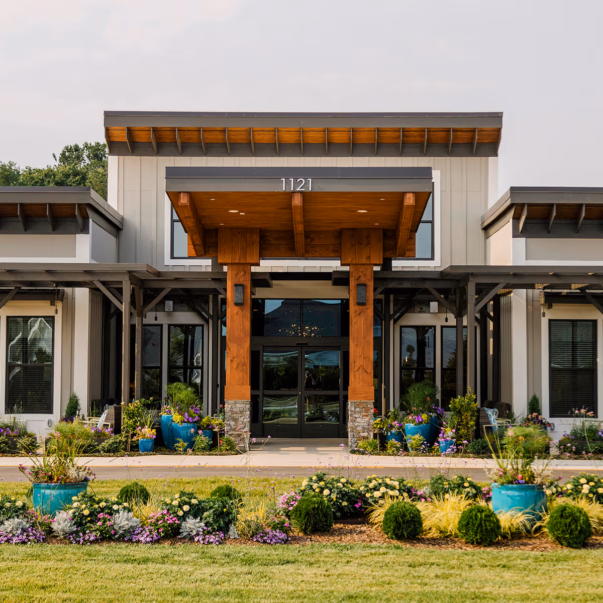Front exterior view of a modern senior living facility building with a covered entrance supported by wooden beams. The building is surrounded by well-maintained landscaping with colorful flowers and green shrubs in blue pots and garden beds.