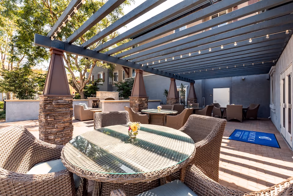 Outdoor covered patio with wicker tables and chairs under a pergola with string lights at a senior living facility.