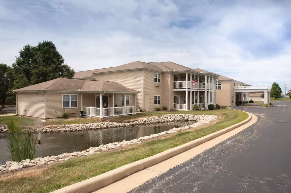 Exterior view of a two-story beige senior living building with balconies, a driveway, and a small pond in the foreground.