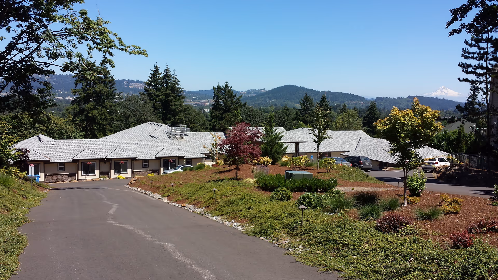 View of Clackamas Heights Senior Living facility from a driveway, showing a single-story building with a gray roof surrounded by landscaped greenery and trees, with hills and a snow-capped mountain visible in the background under a clear blue sky.