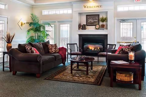 A cozy living room with two dark brown sofas adorned with patterned cushions, a small round wooden coffee table on a decorative rug, and a fireplace with a 'Welcome' sign above it. The room has large windows and glass doors letting in natural light, with plants and decorative items on side tables and the mantel.