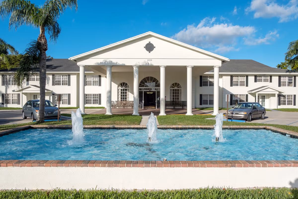White two-story senior living building with columns, fountain, and palm trees in front.