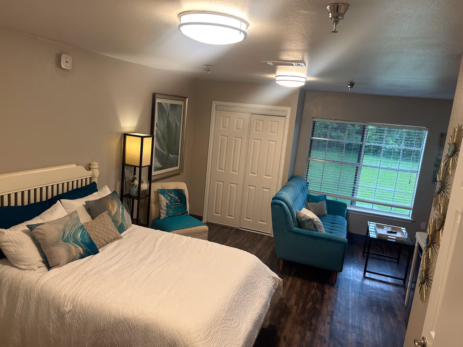A cozy bedroom with a white bed adorned with multiple pillows in shades of teal and beige. To the left of the bed is a tall floor lamp with a square shade and a small shelf holding decorative items. Next to the lamp is a teal cushioned chair with a matching pillow. Across from the bed is a teal sofa with several pillows, positioned in front of a large window with white blinds. A small coffee table sits in front of the sofa. The room has wooden flooring, neutral-colored walls, and two ceiling lights.
