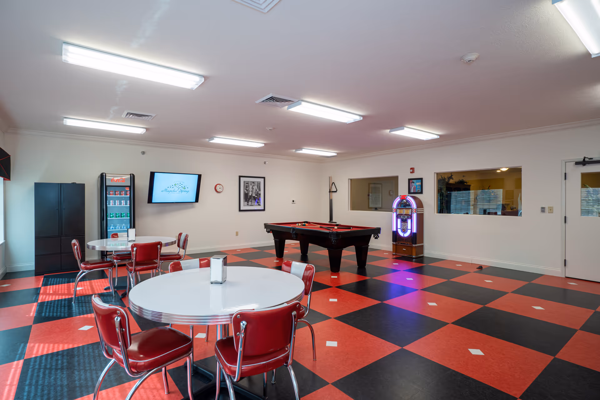 A recreational room with a red and black checkered floor, featuring a pool table, a jukebox, round tables with red chairs, a Coca-Cola vending machine, and a wall-mounted TV displaying the Magnolia Springs logo.